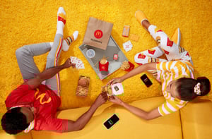 A young man and woman dressed in McDonalds branded clothing are eating their McDelivery order on a yellow couch